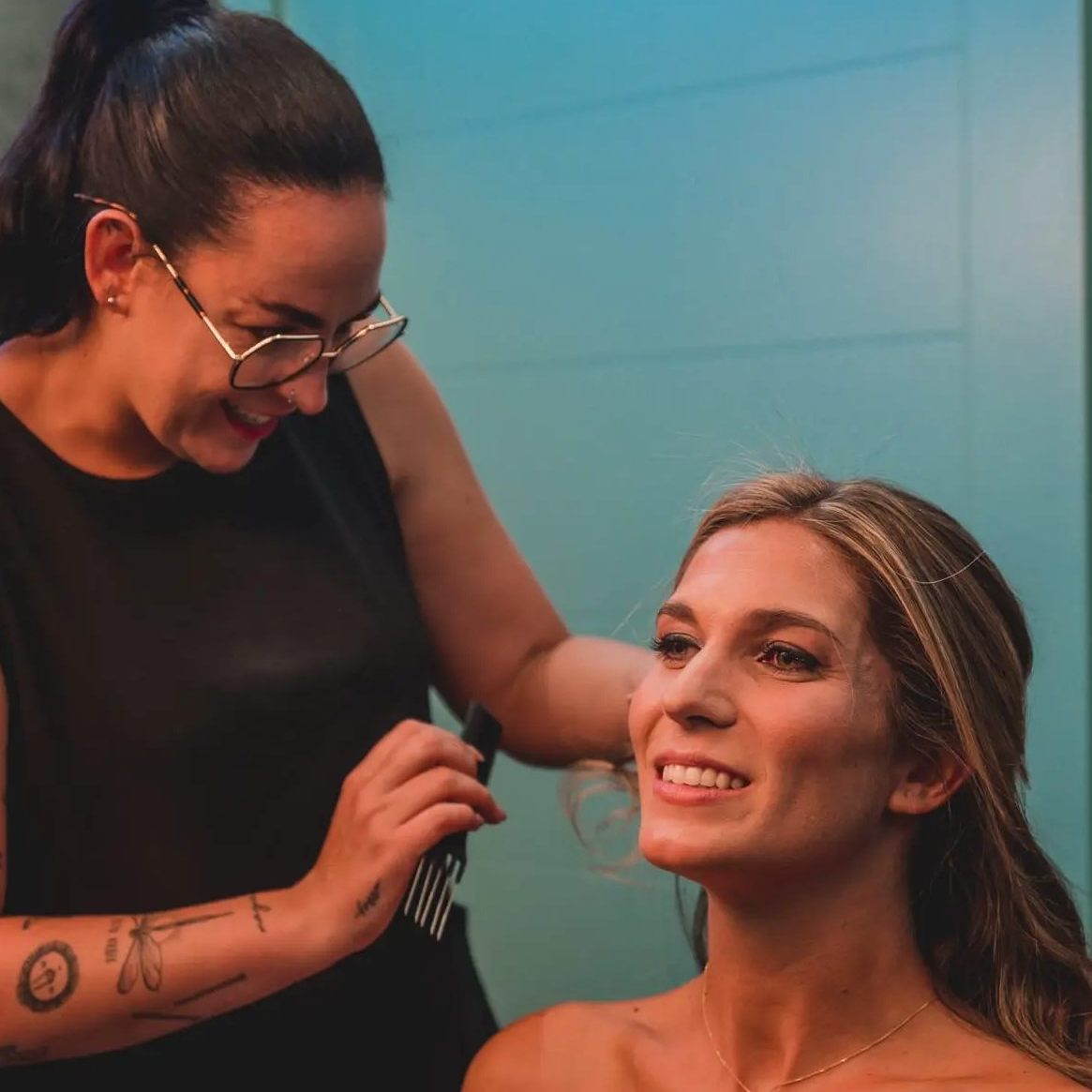 Estilista profesional trabajando en el peinado de una mujer rubia, con una gran sonrisa, durante una sesión de preparación para una boda.